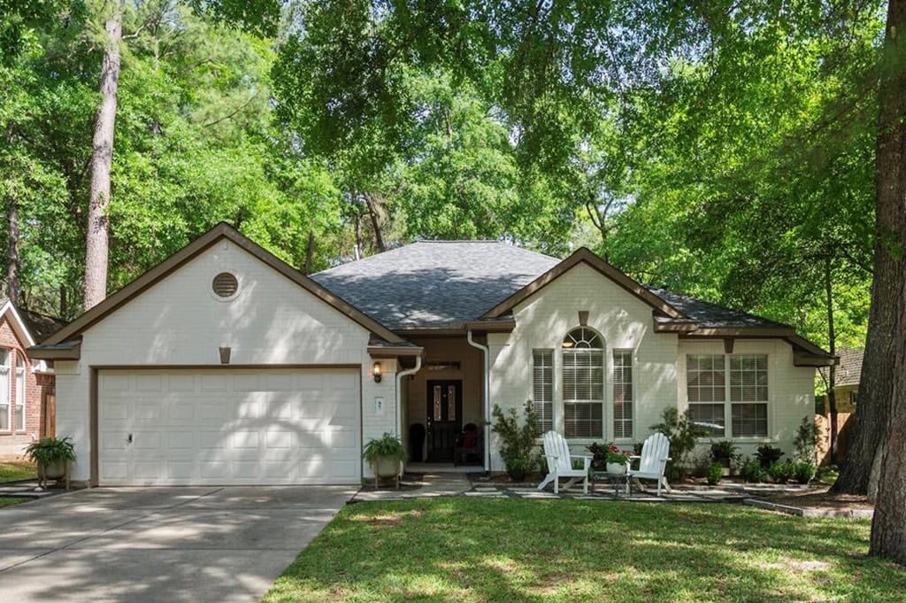 Cozy suburban house with a well-maintained front yard, surrounded by tall green trees, featuring a two-car garage, white siding, and charming outdoor seating area with white chairs and potted plants.
