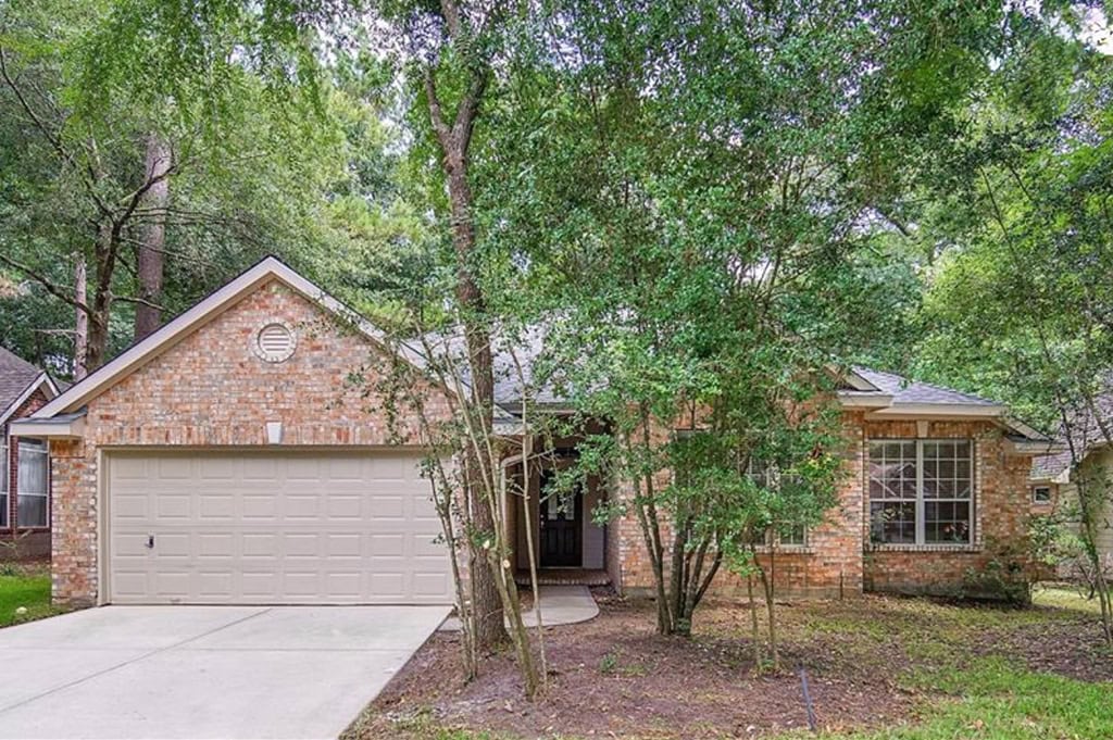 Lush green trees surround a cozy brick house with a two-car garage, creating a serene and inviting outdoor space. The house features large windows, adding charm and natural light.