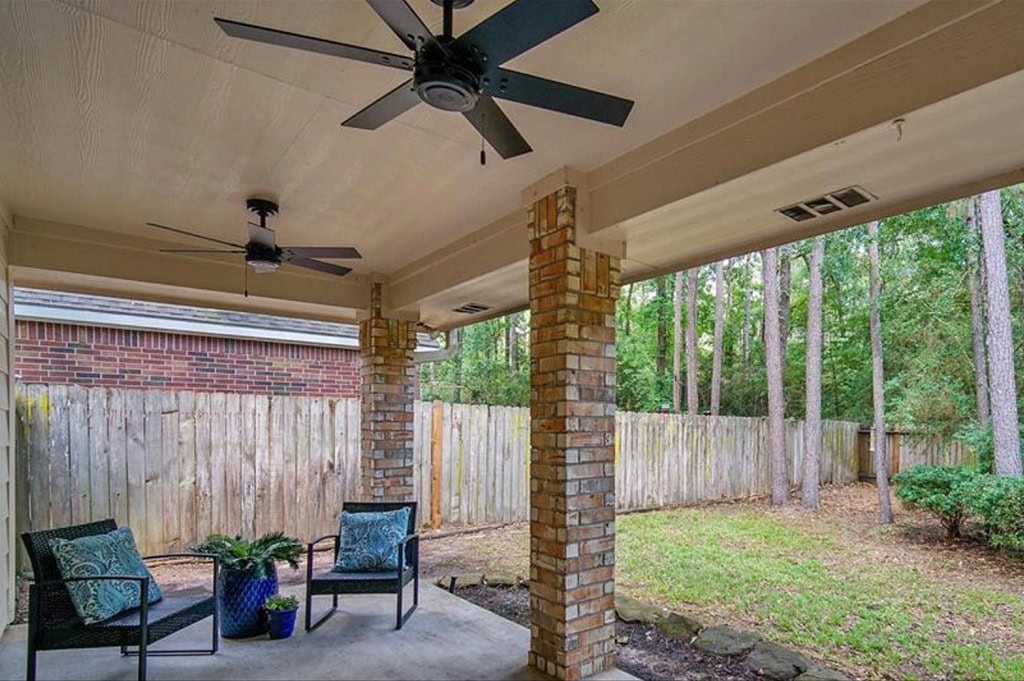 Cozy covered patio with brick columns, ceiling fans, and outdoor seating in a backyard enclosed by a wooden fence, surrounded by lush green trees.