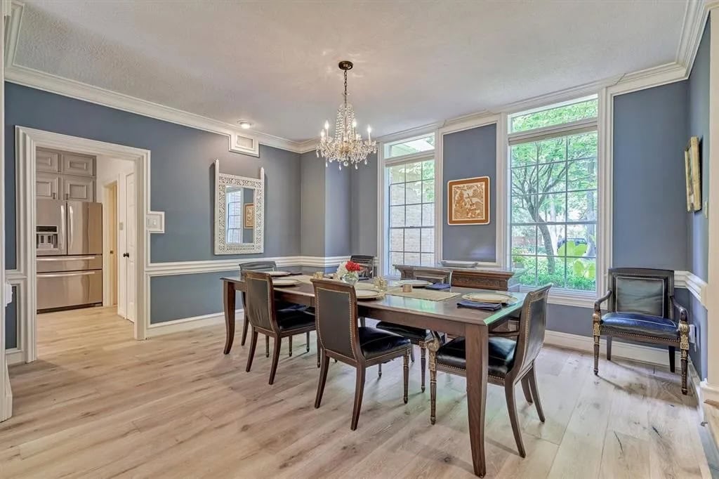 Elegant dining room with blue walls, white trim, large windows, and a crystal chandelier, featuring a wooden dining table with black leather chairs and classic decor elements.