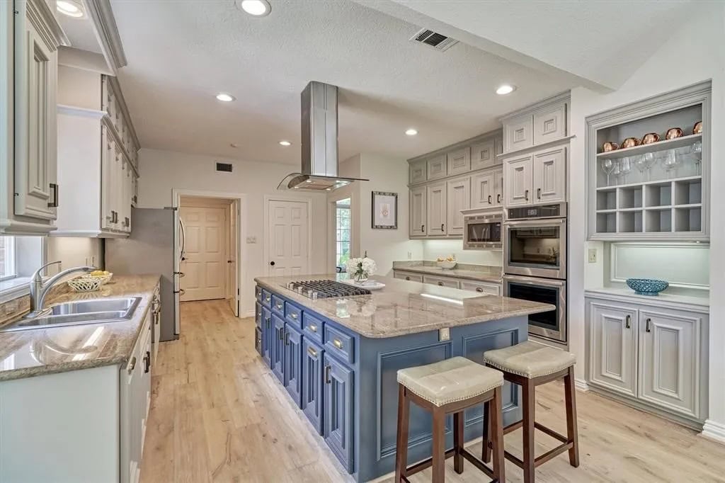 Bright and spacious kitchen with a central island, modern appliances, and ample cabinetry in light gray and blue tones. Natural light floods the space through multiple windows, highlighting the elegant design.