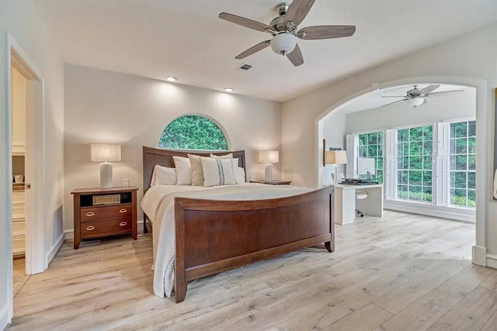 Modern bedroom with wooden bed frame and nightstands, soft lighting, ceiling fans, and large bay window overlooking lush greenery, creating a cozy and airy retreat interior design.